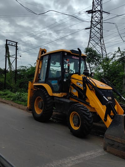 An unoccupied, yellow JCB backhoe loader is parked on the side of an asphalt road. The vehicle faces the viewer's right, with its front loader down and its rear bucket raised. The backhoe is positioned on a slight incline, with the right side of the vehicle closer to the road's edge and dense green foliage. Overhead, a large electricity pylon with numerous wires is visible against a cloudy, overcast sky. In the background, to the left of the pylon, a smaller utility pole with vines growing on it is also present. The scene appears to be in a semi-urban or rural area, possibly on the outskirts of Malkapur, India, given the presence of agricultural and electrical infrastructure. There are no people visible in the image.