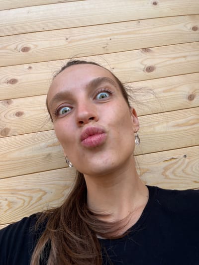 A young woman with brown hair pulled back in a ponytail is pictured close-up against a background of horizontal wooden planks. She is wearing a black t-shirt and clear, teardrop-shaped earrings. Her eyes are wide and her lips are pursed as if she is about to kiss. The image appears to have been taken outdoors, possibly on a balcony or patio, given the visible wood grain of the structure. There is no information about the location being in Bistrica, Montenegro.