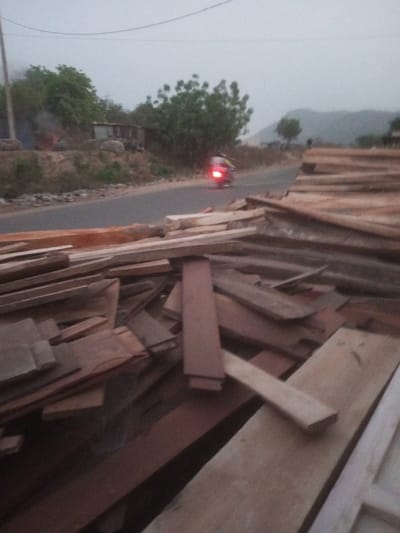 This image captures a scene in Ebiya, Nigeria, under an overcast sky. In the immediate foreground, a substantial, uneven pile of various lengths and widths of sawn timber planks dominates the lower half of the frame. The timber displays shades of light brown and reddish-brown, indicative of different wood types and weathering. Behind this pile, a paved road extends horizontally across the midground. A blurred motorcycle, its red taillight illuminated, is visible traveling from right to left on this road. To the left of the road, an elevated, unpaved embankment supports sparse green foliage and a small structure with a light-colored roof, possibly corrugated metal, from which a plume of light smoke rises. A utility pole is partially visible on the far left. On the right side of the road, further stacks of timber are visible extending into the background. Distant hills are faintly discernible under the hazy sky.