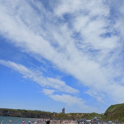 This image depicts a crowded beach scene in Ballybunnion, Ireland, under a sky filled with scattered white clouds. A rocky cliff face with a medieval tower, known as Ballybunnion Castle, is visible on the left side of the frame, overlooking the sea. The beach is populated with numerous individuals enjoying the water and the sand. Some people are swimming, while others are wading near the shore. Beach umbrellas and tents are set up on the sand, indicating recreational activities. The water appears calm, with gentle waves reaching the shore. Cars and a van are parked on a road that winds up the grassy hillside on the right side of the image. The overall atmosphere is one of a busy summer day at the beach, with a prominent historical landmark in the background.