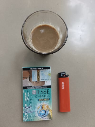 The image shows a top-down view of a glass of coffee, a pack of cigarettes, and an orange lighter placed on a textured surface, possibly a table or floor. The coffee in the glass is light brown and appears to have milk or creamer mixed in. The cigarette pack is teal and white, with text indicating "PERINGATAN" (Warning) and "ESSE CHANGE DOUBLE". The lighter is orange with the brand name "Cricket" printed vertically in white. The overall scene suggests a casual setting, likely indoors. There are no people or specific landmarks visible that would indicate its location within Meunasah Pupu, Indonesia.