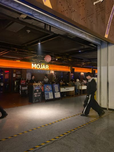 A tilted, long shot captures the exterior of "MOJAR," an establishment lit with bright orange lights. People are seated at tables inside and outside, with various signs and menus visible. A person in black athletic wear walks along a striped gray surface. A pink balloon hangs outside the entrance. The scene conveys an evening ambiance in a public space.