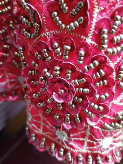 A close-up shot reveals vibrant red fabric intricately adorned with sequins and beads. The sequins, arranged in a circular, floral pattern, shimmer with a translucent quality. Small, gold-colored beads accent the sequins, creating texture and contrast. Gold thread outlines the embellished areas. The overall effect is luxurious and detailed, suggesting a garment or accessory with elaborate craftsmanship.