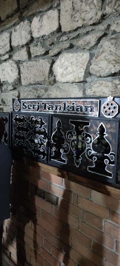 The image shows a sign with the name "Serj Tankian" in raised lettering on a dark background with a white outline, mounted on a stone wall. To the left and right of the sign are decorative pomegranate shapes with cutouts. Below the sign are three dark panels with intricate white scrollwork designs. The sign and panels are mounted on a wall constructed of rough-hewn stones, which transitions into a brick wall in the lower portion of the image. The overall lighting suggests an indoor or dimly lit outdoor environment. The location in Voskevaz, Armenia, is not explicitly visible in the image itself but is provided as context.