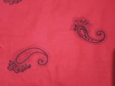 A close-up shot reveals a red fabric adorned with paisley patterns. The paisley motifs are dark gray, adding contrast to the vibrant red background. Two complete paisley figures are easily visible, while a third is partially cut off at the edge of the frame. The fabric appears smooth and slightly textured.