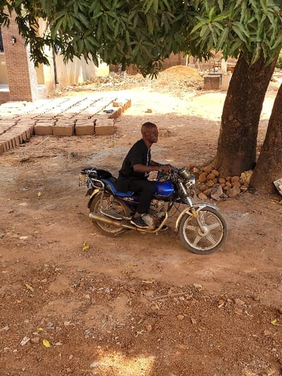 The scene depicts an outdoor area in Ebenebe, Nigeria. A male individual, appearing middle-aged, with short dark hair and dressed in a black t-shirt and black trousers, is seated on a stationary blue and silver motorcycle. He is positioned facing left in the frame, with his hands on the handlebars. The motorcycle has chrome elements, including the front fender and exhaust pipe, and features alloy wheels.
The ground is unpaved, consisting of reddish-brown soil with visible small rocks and dry leaves. To the right of the motorcycle, two tree trunks rise, with dense green foliage extending overhead, providing shade. Small stones are accumulated at the base of the tree trunks.
In the background, to the left of the motorcycle, multiple rows of light brown, rectangular bricks or blocks are neatly stacked on the ground. Beyond these, a partially constructed building with exposed reddish-brown brick walls and light-colored window frames is visible. To the right background, an earthen mound and a concrete structure, possibly a well or foundation, are present. The overall environment suggests a construction or residential area.