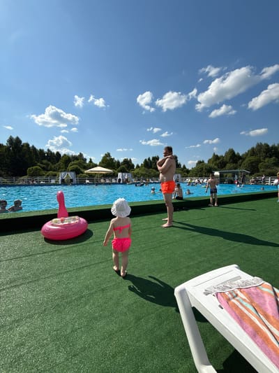 This is a daytime, eye-level, outdoor, full shot photograph taken at a pool facility. The scene depicts a bright, sunny day with a clear blue sky dotted with fluffy white clouds. The foreground is dominated by artificial green turf, upon which a white plastic lounge chair with a striped towel draped over it is visible on the right side. To the left of the chair, a young child wearing a white sun hat and a bright pink bikini is walking away from the camera. In front of the child, a large, inflatable pink flamingo pool float rests on the turf. Beyond the turf is a large, blue swimming pool filled with people. In the background, beyond the pool, is a line of trees and a white fence, suggesting a natural, possibly rural, setting. A few white gazebos with draped curtains and a large cream-colored umbrella are set up near the pool. People are seen swimming, lounging, and socializing in and around the pool. The overall atmosphere is one of leisure and recreation. The location is identified as Koloksha, Russia.