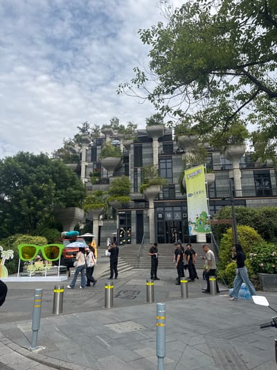 This image captures a street view in Putuo District, China, featuring a modern building with a unique architectural design. The building's facade is adorned with numerous protruding planters, creating a tiered, organic look. Trees and greenery are integrated into the building's structure, cascading down from the elevated platforms.

In the foreground, a large, bright green novelty pair of sunglasses is displayed on a white stand, accompanied by a palm tree graphic and text in an Asian language. An orange SUV, possibly part of a display or advertisement, is parked behind the sunglasses, adorned with surfboards and an umbrella. Several people are present in the scene, some walking and others standing near the building's entrance. A yellow banner with text and graphics is prominently displayed on a pole near the building entrance. The ground is paved with grey interlocking tiles, and metal bollards are placed along the walkway. The sky above is overcast with scattered clouds. The overall impression is a vibrant, contemporary urban environment with an emphasis on greenery and modern design.