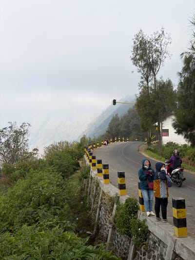 In Ngadas, Indonesia, two women stand on a concrete wall next to a winding road. The woman on the left wears a blue hooded jacket and jeans, holding a red bag and a small item. The woman on the right wears a multi-colored jacket and black pants, holding a phone. Behind them, a red and white motorcycle is parked, and a person in a purple jacket is seated on it. The road is bordered by black and yellow striped bollards. In the background, dense green foliage lines a retaining wall, and a misty, mountainous landscape stretches into the distance. A traffic light hangs over the road.