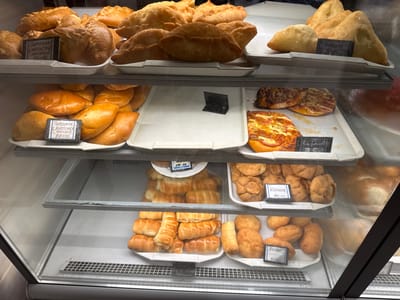 The image shows a refrigerated display case filled with various baked goods, likely in a bakery or shop in Inzer, Russia. The items are arranged on multiple shelves.

On the top shelf, there are several golden-brown pastries, some crescent-shaped and others oval. One sign reads "ПИРОЖОК С КАПУСТОЙ ПЕЧЕНЫЙ 50-00," indicating cabbage pies priced at 50 rubles.

The second shelf contains more golden-brown baked goods, including several oval-shaped pies, some appearing to have a filling. A sign with "ПИРОЖОК С КАРТОФЕЛЕМ ПЕЧЕНЫЙ 50-00" indicates potato pies also priced at 50 rubles. There are also some pizzas or flatbreads with toppings on this shelf.

The third shelf holds a large quantity of small, elongated pastries, some appearing to be filled rolls. There are also some round, fried pastries. Signs on this shelf read "КУРНИК 90-00" for chicken pies at 90 rubles, and "БЕЛЯШ 90-00" for Belyash pastries at 90 rubles.

The bottom shelf appears to be mostly empty with a metal grate at the bottom, possibly for ventilation.

The display case is made of glass and metal, with bright interior lighting. No people are visible in the image. The overall impression is of a well-stocked food establishment.