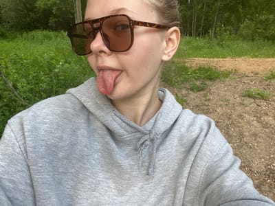 This is a close-up, upside-down selfie of a person with fair skin. The person is wearing a gray hooded sweatshirt with drawstrings and a silver necklace. They are also wearing brown, tortoise-shell patterned sunglasses. The person's tongue is sticking out.
The background appears to be an outdoor scene with greenery, suggesting a natural setting like a park or forest. There's also a sandy path visible. The lighting suggests it might be daytime, and the greenery implies a temperate season. The overall mood conveyed by the image is playful and candid. No text is visible in the image. The provided location context is Klepiki, Russia.