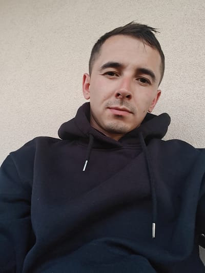 In Shkurupiivka, Ukraine, a young man with short dark hair and a slight stubble is captured in a close-up, outdoor selfie. He is wearing a black hooded sweatshirt. The background consists of a textured, light-colored wall. The man's head is tilted slightly, and he is looking directly at the camera with a neutral expression. The lighting suggests it is daytime.