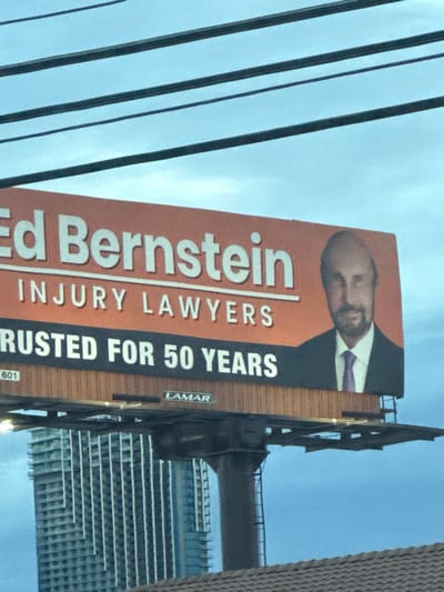 A large billboard is prominently displayed against a light blue sky with faint cloud cover in Paradise, United States. The billboard features an orange upper section with white text reading "Ed Bernstein INJURY LAWYERS" and a black lower section with white text stating "TRUSTED FOR 50 YEARS." To the right, a photograph of a balding man with a beard, wearing a dark suit and a purple tie, is integrated into the design. The "LAMAR" logo is visible on the billboard's support structure, along with the number "601" on the lower left corner of the advertisement panel. Multiple black power lines traverse horizontally across the upper portion of the frame. In the background, a tall, multi-story building with a vertical facade is partially visible. The lower foreground shows a section of a dark-colored rooftop.