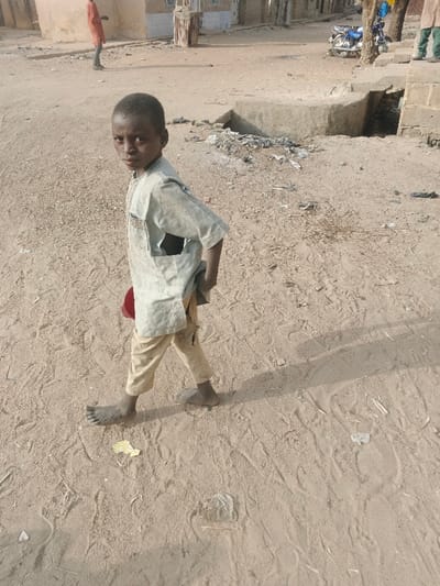 The image depicts an outdoor scene on an unpaved, sandy street in Kwachiri, Nigeria, under clear daylight conditions, indicated by strong shadows.
In the foreground, a young male, appearing to be a child between 6 and 10 years old, stands facing the camera with a direct gaze. He has dark skin and short, dark hair. He wears a light-colored, possibly off-white or pale green, long-sleeved tunic (qameez style) with visible embroidery near the neckline and a tear on the left side, exposing a darker layer or his skin underneath. His lower attire consists of light brown or khaki-colored trousers that extend to his mid-calves. He is barefoot, and his feet show dirt accumulation. He holds a small, red, bowl-shaped object in his right hand, positioned near his hip.
The ground consists of light brown sand and dirt, exhibiting numerous footprints and scattered small pieces of litter, including a yellow fragment of paper or plastic near the child's left foot and a clear plastic item further back. A shadow from the child extends to his right.
In the mid-ground, a concrete curb or drainage structure runs horizontally. Adjacent to it, a collection of refuse and debris is visible. Beyond this, several single-story, light-colored buildings with windows and doorways line what appears to be a street. These structures appear to be constructed from local materials and show signs of wear.
In the background, on the far left, an individual dressed in an orange or reddish garment stands facing away from the camera. Further to the right, behind the concrete structure, a blue motorcycle is parked near a tree. The lower legs of another individual, clad in green trousers, are partially visible near the tree. The overall environment suggests a low-income urban or semi-urban area.