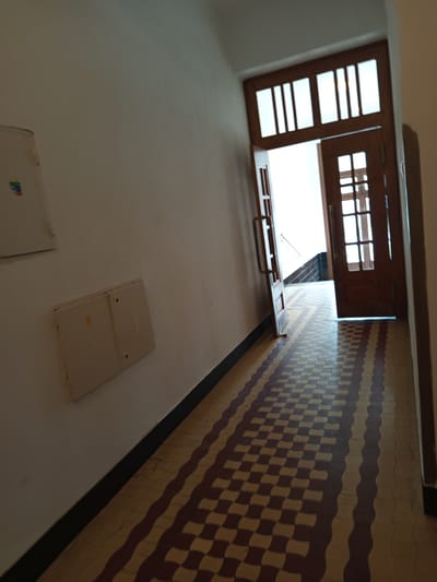 The image shows a view looking up into an open window and hallway in what appears to be an apartment building or similar structure. The subjects are architectural elements and interior details.
In the foreground, a white wall takes up a significant portion of the lower half of the image, with a light beige electrical box mounted on it. Another similar box is partially visible in the bottom left corner, with a colorful logo. The wall transitions to a tiled floor in the background.
The focal point of the image is an open wooden-framed window, with its upper sash tilted open. The window has a grid pattern in its upper portion. Below the window, a ledge or sill is visible with a metal handle. Beyond the window, a bright white light suggests it is daytime, possibly with bright sunlight outside.
To the right of the window, a hallway wall is covered in patterned tiles. The tiles are arranged in a wavy, interlocking pattern, with alternating colors of beige and dark red or brown. The lighting creates distinct shadows on the tiled wall, indicating a light source from the window.
The scene suggests an interior residential or communal space. There is no indication of people or specific activities, other than the passive presence of the architectural features. The overall impression is one of an interior view looking outwards and upwards. No visible text is discernible in the image, except for a very small, unreadable logo on the electrical box in the lower left.