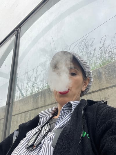 A medium shot shows a person exhaling a large puff of vapor in what appears to be a bus shelter in Mensdorf, Luxembourg. The subject, possibly female, is wearing a light-colored hat with a decorative rim, a black fleece jacket with green embroidered lettering, and a striped shirt. Glasses are hooked on the shirt. The backdrop includes a cloudy sky and some trees visible through the shelter's glass panels and metal framing.