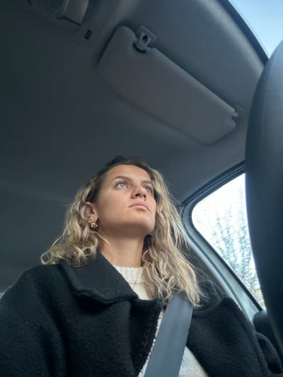 A woman with curly blonde hair is seated in a car, wearing a black jacket and a white sweater. She is wearing a seatbelt and golden earrings. The car's visor is visible above her to the left. She is looking up toward the car window, which shows a glimpse of the sky and bare trees. The angle is from below, looking up at the woman in the car.