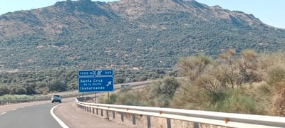 The image captures a scenic view of a highway in Spain, likely in the vicinity of Santa Cruz de la Sierra, given the signage. In the foreground, a paved highway curves to the left, bordered by a white line on the left and a guardrail on the right. A single car, a silver hatchback, is visible on the highway, traveling away from the viewer.
A blue road sign is prominently displayed on the right side of the highway. The sign contains several pieces of information: "1000 m" (1000 meters), a symbol indicating a fork in the road or a junction, and the number "268". Below this, it reads "Santa Cruz de la Sierra" and "Ibahernando", with an arrow pointing to the right, indicating directions to these locations.
The background is dominated by a large, rocky mountain covered in dense vegetation. The vegetation appears to be a mix of shrubs and trees, creating a textured, green-grey blanket across the mountainside. The sky is clear and blue. The overall impression is of a rural, mountainous landscape with a well-maintained highway facilitating travel.