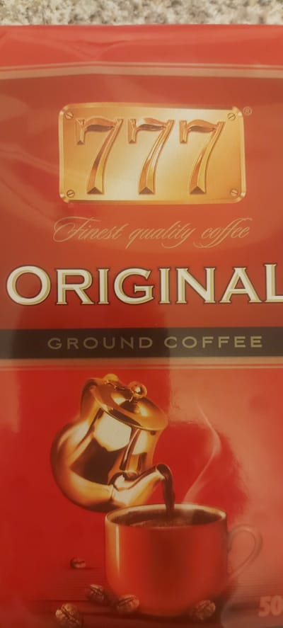 A close-up, vertical shot of a red coffee package. The top of the package features a gold embossed logo with three sevens. Below the logo, in elegant script, are the words "Finest quality coffee". Further down, large, bold white letters spell out "ORIGINAL". A dark horizontal stripe with the text "GROUND COFFEE" in white caps runs below the word "ORIGINAL". The bottom of the package depicts a golden coffee pot pouring coffee into a red mug. Several coffee beans are scattered on a dark surface in the foreground. The image is a product shot, likely taken in a studio or retail environment, and is not set in a specific outdoor location. There are no people in the image. The image is not located in Kohtla-Järve linn, Estonia.