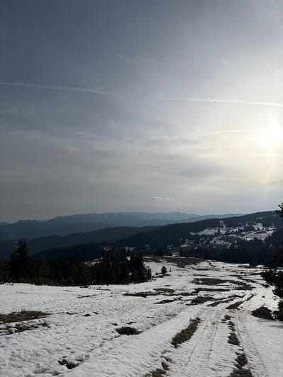 The image depicts a winter landscape in Momchilovtsi, Bulgaria, with snow-covered terrain. A mountain range is visible in the background, with trees and snow patches. The sky is bright with clouds and a contrail. The foreground shows a snowy path with tracks.