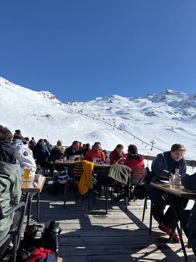 A group of people are sitting at tables on a deck, located on a mountainside ski slope. There are empty beer glasses on the tables. The people are dressed in warm winter clothing, and some are wearing ski helmets. There are multiple tables filled with people, facing the skiers and snow-covered slopes. The sky is blue and clear. The scene is located in the Les Belleville ski resort in France. The coordinates provided are for the resort's general location. The exact location of the scene is not evident.