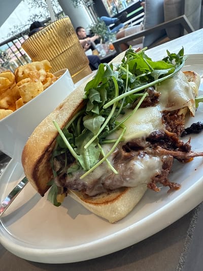 A close-up, slightly rotated shot shows a sandwich on a white oblong plate alongside a white rectangular dish of waffle fries, likely taken at a restaurant in Oak Brook, Illinois, USA. The sandwich is composed of a toasted roll filled with shredded meat covered with melted white cheese and topped with fresh arugula. The waffle fries are piled high in the small dish. The plate sits on a shiny tabletop. In the background, out-of-focus, there is a view of people and the restaurant interior through a window. There is a gold-colored corrugated object on the tabletop. The shot seems to have been taken indoors with available light.