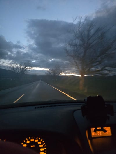 The image shows a view from inside a car driving on a road, likely a highway, at dusk or dawn. The sky is overcast with heavy clouds. Bare trees line the roadside. The road is asphalt with a yellow line marking the lanes. The dashboard of the car is visible, including a digital display reading "01. 10." and a portion of the speedometer with orange illumination. A phone mount is attached to the dashboard. The geographical location of Marino Pole, Bulgaria, is not determinable from the image.