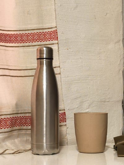 This is a still life photograph featuring a stainless steel thermos and a ceramic cup placed on a white surface, likely a tabletop. In the background, a textured, off-white wall is visible on the right, while a cream-colored textile with red and brown horizontal striped patterns is draped on the left. The lighting is soft, casting subtle shadows. There are no people or animals present in the image. The objects are positioned in a simple, uncluttered arrangement. The image does not provide any information that would indicate its location in Mikkeli, Finland.
