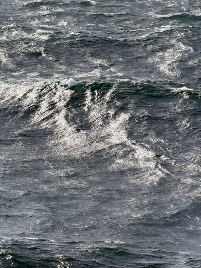 The image displays a rough, choppy sea under bright sunlight, with waves reflecting the light. The water is a dark, deep blue-green, with white foam cresting on the wave tops. The texture of the water is dynamic and uneven, suggesting strong winds and currents. The overall impression is one of a wild and powerful natural environment. This image does not depict any specific location in Strzelno, Poland, as Strzelno is a landlocked town.