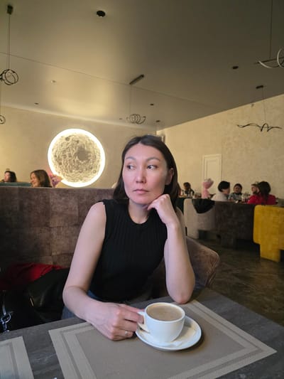 A woman sits at a table with a cup of coffee. Other people are visible in the background. The scene is likely inside a cafe or restaurant. The location is Lensk, Russia.