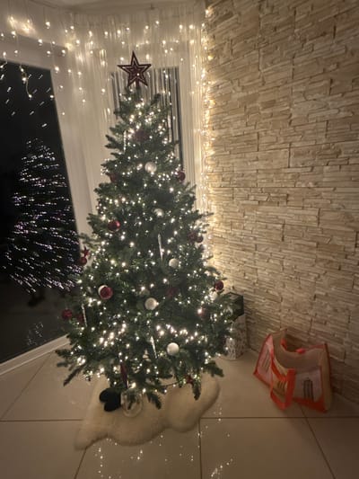 The image displays an inviting indoor scene, likely within a home in Wipperfürth, Germany, decorated for the Christmas season. The primary subject is a large, vibrant Christmas tree, lavishly illuminated with numerous small white LED lights. The tree is further adorned with spherical ornaments in red and silver-white, and a distinctive red star serves as its topper.

The tree is situated in front of a window, which reflects the bright indoor lights, creating a dazzling visual effect. The darkness visible through the window suggests that the photograph was taken at night or during the late evening.

In the background, a striking wall features light-colored, stacked stone or brick veneer, adding a rustic yet modern texture to the space. Additional strings of white fairy lights are draped along the wall, emphasizing the festive atmosphere. To the right of the tree, a fluffy white or cream-colored rug is partially visible on a light-toned floor. A wrapped gift box can be seen tucked behind the tree, and a patterned fabric bag or basket rests in the upper right corner of the frame.

The overall scene conveys a sense of warmth, coziness, and holiday cheer. No people are visible in the image, and there is no discernible text. The focus is entirely on the festive decorations and the inviting ambiance of the room.