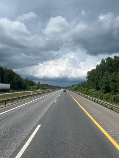 The image depicts a highway in a rural setting, with a dark, cloudy sky overhead. The road has multiple lanes, and there is a guardrail on either side. Dense green trees line the highway. A few cars and a tanker truck are visible on the road. The location in Kiyasovo, Russia, is consistent with a highway traversing a forested area. The scene appears to be captured from a moving vehicle.
