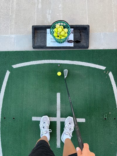 An overhead shot shows a pair of white athletic shoes standing on a green turf golf practice mat. The person wearing the shoes is holding a black golf club with a silver clubhead. Two yellow golf balls and a small blue object are on the mat in front of the person. A green basket filled with yellow golf balls sits on a grey mat to the upper left. A pair of dark sunglasses rests next to the basket. The practice mat is bordered by a white line and a light-colored tiled area. This scene is at a golf driving range in Tsada, Cyprus.