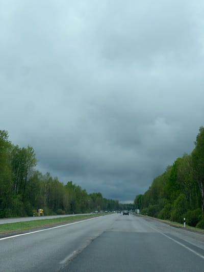 A photograph depicts a grey, overcast sky above a road lined with trees in Глівінскі сельскі Савет, Belarus. The road is wet, suggesting recent rain, and several vehicles are visible in the distance. Trees flank both sides of the road, and a sign is present on the left side. The sky is heavily clouded.