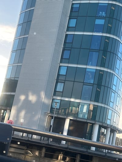 The image displays a multi-story modern building identified as Pier Quays at Compass Point, Gillingham Marina, Gillingham, Kent, United Kingdom. The building features a distinctive curvilinear façade composed primarily of blue-tinted reflective glass panels, alternating with a prominent vertical section clad in horizontal grey metallic louvered panels. Intense sunlight illuminates the structure from the left, creating bright reflections across both glass and metal surfaces.
On a mid-level floor within one of the curved glass sections, a human head and upper torso are visible through a window, positioned near interior furniture resembling chairs and a desk. Other windows reveal internal light fixtures, curtains, or indistinct objects suggesting residential spaces.
The ground floor of the building is supported by several white cylindrical pillars, with dark, possibly shuttered, entryways. A dark metallic handrail extends horizontally across the lower foreground, partially obscuring a paved platform or dock area below the building. A lamppost stands near the building's base. In the far background, to the right, several boat masts are faintly discernible, indicating the building's proximity to a marina. The sky above is partially cloudy and light blue.