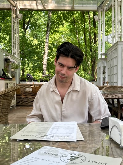 A medium shot depicts a dark-haired man in a light-colored, partially unbuttoned shirt seated at an outdoor table. He is gazing towards the right, seemingly focused on a menu with Cyrillic script. The menu is placed on the table, with the words "КУХНИ" (Kitchens) visible. A second menu lies to the right, showcasing "МЕНЮ ЗАКУСКИ И САЛАТЫ."

The setting seems to be a restaurant in Arkhangel’skoye, Russia. The scene is filled with abundant natural light. Dense foliage and trees are visible in the background, suggesting a park or garden setting. The outdoor seating area has woven wicker chairs and the table is made of glass with a pattern visible underneath. The overhead structure appears to be a transparent roof, which allows more natural light. Napkins are present on the table.