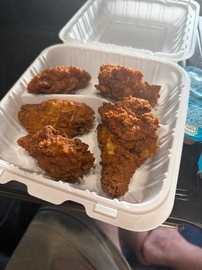 A white takeout container holds five pieces of fried chicken. The container is open, and its lid is propped up behind the chicken. The chicken pieces are golden brown and appear crispy. The container sits on a dark surface, possibly a table. Two small blue packets of ranch dressing are visible to the right of the container. The lower portion of the image shows the legs of a person. The overall scene suggests a casual meal. Raphine, United States is the stated location.