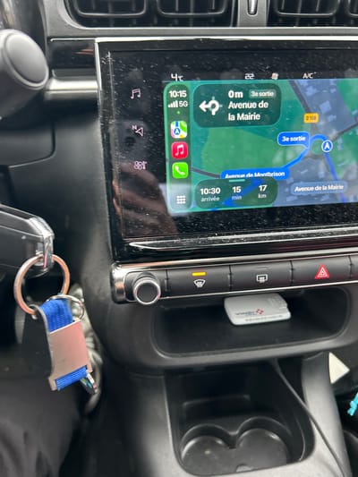 The image depicts the interior of a vehicle's dashboard, focused on the central infotainment system and surrounding controls. A dusty digital display presents a navigation interface, indicating a current time of 10:15 with full 5G cellular signal and an external temperature of 4°C. The navigation shows a route with the next instruction being "0m 3e sortie Avenue de la Mairie," leading to an estimated arrival at 10:30 after 15 minutes and 17 kilometers. Prominent street names visible on the map include "Avenue de Montbrison" and "Avenue de la Mairie," placing the vehicle's navigation within the city of Saint-Cyprien, France, specifically the one located in the Loire department, due to the proximity of Montbrison. Below the screen, physical controls are present: a rotary knob to the left, and a row of buttons including an illuminated front windshield defroster, a rear defroster, and a red hazard warning light button. A storage cubby beneath these controls contains a white "VINCI Autoroutes" toll transponder. To the left of the center console, a set of car keys with a blue fabric lanyard and a metallic keychain is visible. Reflections of the vehicle's interior and an unseen person, likely holding a device, are discernible on the glossy surface of the screen.