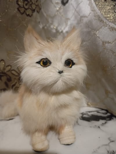 A close-up, frontal shot shows a fluffy, light-colored stuffed animal resembling a fox or cat. Its fur is light beige with subtle orange-brown patches, and it has large, round, amber-colored eyes with black pupils, giving it an alert and curious expression. The creature has pointed ears and a small black nose and mouth. Its body is rounded and appears to be made of soft, plush material. The stuffed animal is sitting on a flat surface that has a marbled pattern of black and white. Behind the creature, there's a textured, possibly fabric, background with a subtle floral or ornate pattern in muted brown and gold tones, suggesting an indoor setting. The lighting is soft and diffused, highlighting the texture of the fur and the reflective quality of the eyes. There is no visible text in the image.