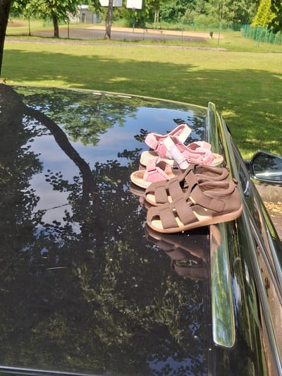 The image shows two pairs of children's sandals placed on the dark, reflective hood of a car. One pair is pink, with a light pink sole and light grey straps. The other pair is brown, with tan soles and brown straps. The sandals are positioned towards the right side of the hood, with the brown sandals in front of the pink ones. The car's hood reflects the surrounding environment, including trees with green leaves and the blue sky. In the background, a grassy area with trees and a fence is visible, suggesting an outdoor setting, possibly a park or a residential yard. The location is Lēdmane, Latvia.