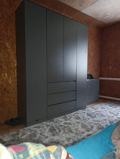 The image shows the interior of a room, likely a bedroom or storage area. A large, dark grey wardrobe dominates the left side of the frame. It has multiple doors and drawers. To the right of the wardrobe is a smaller, matching chest of drawers with several drawers. A black electronic device, possibly a landline phone, sits on top of the chest of drawers, with a thin wire extending from it. The walls are covered with oriented strand board (OSB) panels, a common building material. The floor is covered with a patterned rug, and a blue blanket is partially visible in the foreground. A pair of dark slippers rests on the floor near the base of the wardrobe. The lighting suggests an indoor setting, possibly with artificial light. There are no people visible in the image. The overall impression is of a functional, somewhat unfinished living space. There is no indication in the image to suggest a specific location within Shumerlya, Russia.