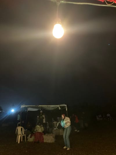 The image depicts an outdoor nighttime scene located in Murera ward, Kenya. A single illuminated incandescent light bulb hangs centrally in the upper frame, serving as a primary light source and emitting a strong glare.

The ground surface is dark, uneven earth. In the right-center foreground, an individual stands, facing slightly left. This person wears blue jeans, a light-colored patterned long-sleeved shirt, and a red beanie. They are holding an object in front of them that emits a distinct light blue-green glow.

To the left, under a temporary white fabric canopy supported by poles, several indistinct figures are gathered, some seated on white plastic chairs. Beneath this canopy, on the ground, are multiple rectangular bales, consistent with hay or straw, and a red plaid fabric item. An empty white plastic chair is positioned to the left of these bales.

Further in the dark background, to the right of the canopy, additional indistinct individuals are visible, some seated on white plastic chairs. Other artificial light sources are faintly visible in the far left background. The overall environment is dark, indicative of night.