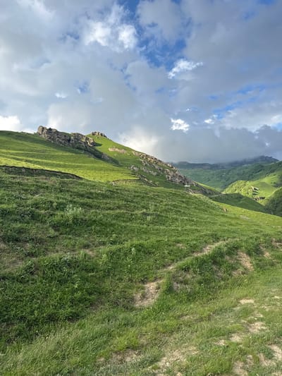 A vast, rolling green mountain landscape is captured under a dramatic sky filled with puffy white and gray clouds. The foreground is dominated by a grassy slope with visible tire tracks or worn paths leading up and across it. In the mid-ground, a steeper, lush green mountainside rises, dotted with rocky outcrops and patches of exposed earth. Further in the distance, more mountainous terrain unfolds, with valleys carved into the landscape, some of which are shrouded in mist or cloud cover, creating a sense of depth and scale. The light suggests it is either early morning or late afternoon, with shadows cast across the slopes. The scene appears to be in a natural, undeveloped area, likely a remote mountain region in or near Верхняя Мара, Russia, given the terrain and vegetation. There are no people or animals immediately visible in the foreground or mid-ground, though a small, reddish-brown shape, possibly an animal, can be seen grazing in the distance on the right side of the image. A signpost is visible on a road or path in the mid-distance on the left.