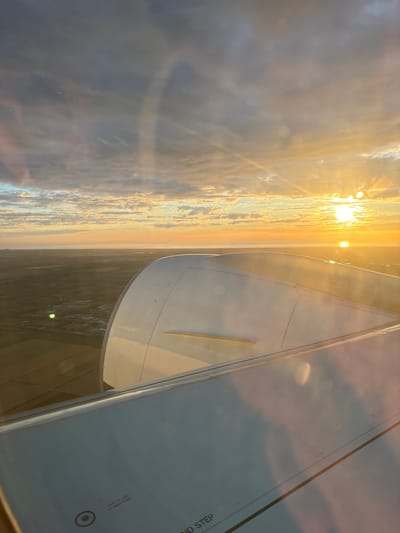 The image captures a view from an airplane window, likely during takeoff or landing at Schiphol Airport (AMS) near Hoofddorp, Netherlands. The setting sun casts a warm, golden light across the scene, illuminating the clouds and the landscape below.

The foreground is dominated by the wing of the airplane, a large, white, metallic structure. The wing's surface is visible, revealing panel lines and some warning labels, including "SAFETY LINE" and "ATTACH POINT." Part of the aircraft's engine nacelle, also white and curved, is visible just behind the wing.

Beyond the wing, the landscape of Hoofddorp unfolds. It appears to be a mix of agricultural fields, characterized by a patchwork of brown and green, and urban areas with buildings and roads. The horizon is flat, suggesting the flat terrain typical of the Netherlands.

The sky is a dramatic display of light and color. The sun is low on the horizon, a bright, intense orb peeking through a band of clouds. Rays of light emanate from the sun, creating a halo effect and casting long shadows. The clouds are a mix of grays and whites, with the undersides illuminated by the sunset, giving them a warm, orange glow. The overall atmosphere is serene and majestic, a common sight during aviation travel at sunrise or sunset.