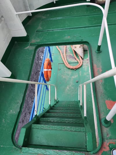 This is a top-down, medium shot of a green metal staircase on the deck of a vessel. The staircase is descending into a rectangular opening, revealing dark, choppy water below, indicating the vessel is in motion. The staircase is flanked by white railings with green bases, which then transition to blue railings closer to the opening. An orange life preserver with white stripes is hanging on the blue railing to the left of the opening. On the deck to the right of the opening, a thick, braided orange rope is coiled loosely next to a metal cleat. The deck itself is painted green and appears weathered with some scuff marks and patches of red. The context of Shal'skii, Russia, suggests this scene likely takes place on a ferry or working boat operating in a port or waterway associated with the city. The lighting is overcast, casting soft shadows.