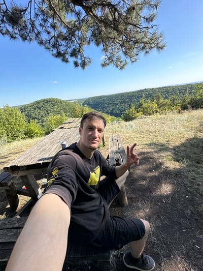 A man is seated on a wooden bench at a picnic table, taking a selfie. He is wearing a black hoodie with a yellow emblem and black shorts. The man is looking at the camera and making a peace sign with his right hand. Behind him, there is a scenic view of rolling green hills and a clear blue sky. The picnic table appears weathered and aged. The surrounding ground is covered with dry grass, dirt, and some scattered debris. The location is identified as being in the city of Nizhyn, Ukraine, likely an outdoor recreation area or viewpoint.