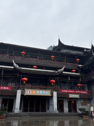 This image displays a multi-story traditional Chinese building, identified as a Tourist Center and Ticket Office, located in Pengshui Miao and Tujia Autonomous County, China. The structure features dark wooden construction with multiple tiered balconies, intricate dark-tiled rooflines with upward-curving eaves, and grey stone elements on the lower facade and pillars flanking the main entrance. Seven spherical red lanterns with yellow tassels are visibly suspended from the balconies and eaves across different levels. Red banners with white Chinese text, including partial messages related to incidents and welcoming the New Year with environmental protection, are strung across the building's facade. The ground in the foreground is dark, wet, and reflective, indicative of recent precipitation. The sky above is uniformly overcast and grey. No individuals are present in the scene. Small potted plants are positioned near the building's entrance.