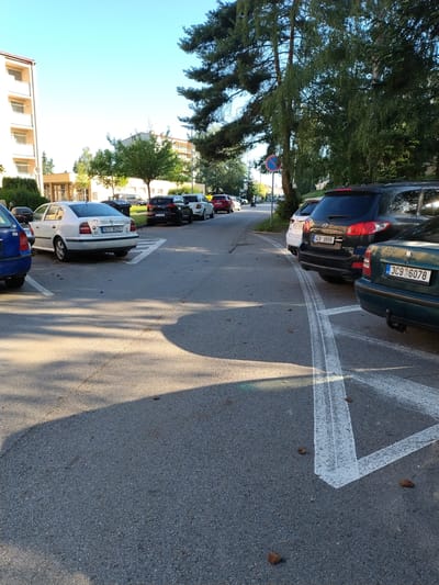 The image shows a parking lot in Písek, Czechia, on a sunny day. Several cars are parked in designated spots, marked by white lines painted on the asphalt. Some of these lines are faded, with tire marks visible on the surface. A few cars are dark-colored, including a black SUV and a dark green station wagon with license plate "3C9 6078". A white sedan with license plate "5K3 8029" is also visible.

The scene is bordered by lush green trees and buildings, suggesting an urban or suburban setting. In the background, residential buildings with light-colored facades and windows are visible. The sun casts long shadows across the parking lot, indicating it is likely late afternoon. There are a few scattered pine cones on the ground. A circular red and blue traffic sign with a white line through it, likely indicating "no stopping," is present near the trees. The overall atmosphere appears calm and ordinary, depicting a typical day in a parking area. No people are clearly visible in the image.