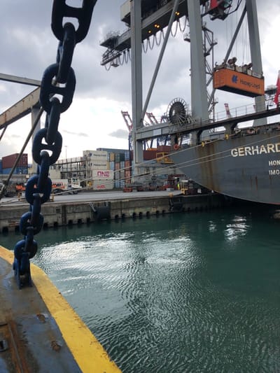 The image depicts a commercial shipping port in La Spezia, Italy, under a cloudy sky. In the immediate foreground, a heavy, dark-colored anchor chain hangs vertically, partially obscured, with a rusted metal surface featuring a prominent yellow-painted edge at the bottom left. The middle ground features calm, dark teal-green water with ripples, bordered by a concrete quay wall equipped with black cylindrical fenders.
To the right, a large grey cargo vessel, identifiable by the partial name "GERHARDT" and "HONG" (likely part of its port of registry, Hong Kong) and "IMO" number on its hull, is moored alongside the quay. Several mooring lines extend from the ship to bollards on the dock.
On the quay behind the ship, numerous shipping containers are stacked, some bearing the logos "Hapag-Lloyd," "ONE," and "MSC." Container handling equipment, including what appears to be a terminal tractor or reach stacker, is visible on the paved dock area. Dominating the background are multiple grey gantry cranes (container cranes), one of which is actively lowering or raising an orange "Hapag-Lloyd" branded container directly above the cargo ship. Additional cranes are visible further in the distance. No individuals are clearly discernible in the scene.