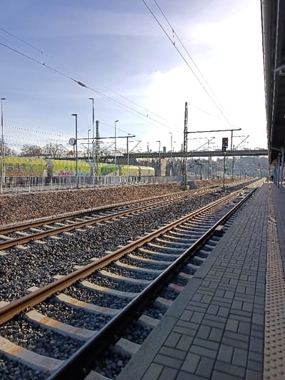 The image presents a railway scene at the Altenburg train station in Altenburg, Germany. In the foreground, a paved train platform occupies the right side, constructed from grey rectangular pavers with a textured tactile warning strip along its edge. Adjacent to the platform, two sets of parallel train tracks are visible, laid on concrete sleepers embedded in dark grey ballast, extending into the distance.
To the left of these tracks, an elevated embankment composed of brown gravel separates them from another set of tracks. On these leftmost tracks, several stationary yellow/lime-green covered freight train wagons are present. These wagons display visible graffiti, including the letters "GATX". A white metal fence or railing runs along the base of the gravel embankment.
Above the tracks, a complex system of overhead electrical lines (catenary) is suspended, supported by multiple metal masts and gantries. Numerous modern-style lamp posts are positioned throughout the scene. A railway signal displaying an amber indication is visible in the mid-ground above the tracks, and a circular clock face is mounted on a pole further to the left. In the background, leafless trees are visible, and an elevated bridge structure crosses the railway lines. The scene is illuminated by sunlight from the right, casting shadows under a clear blue sky with scattered wispy clouds. No individuals are visible within the frame.